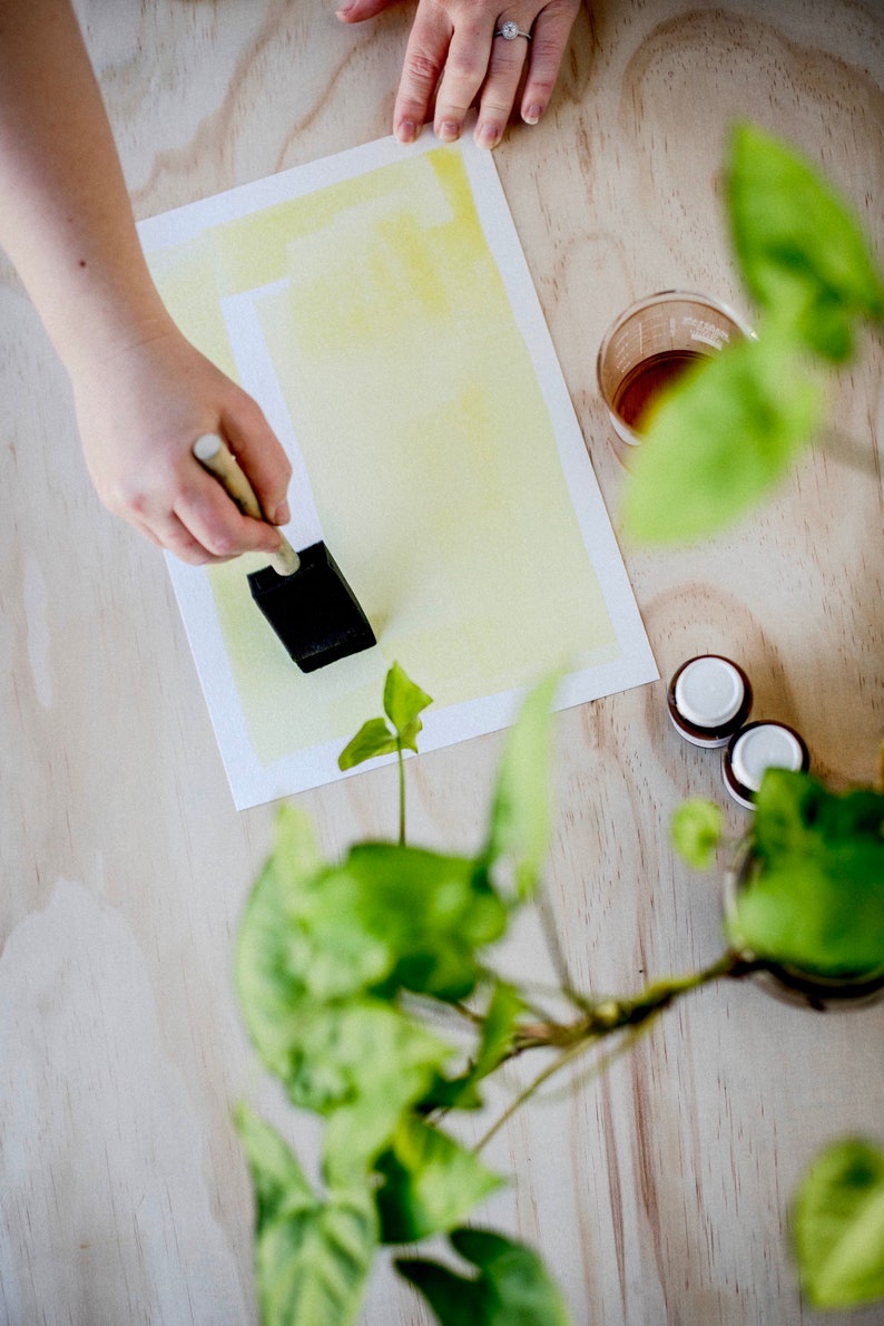 Peut inclure: Une main tenant un pinceau noir au-dessus d'une feuille de papier blanche avec une lavis aquarelle jaune. Le papier est sur une surface en bois clair avec une plante verte en arri&egrave;re-plan.
