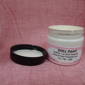 May include: A white jar of doll paint with a black lid next to it. The jar has a pink label with the words "DOLL PAINT" and contact information. The lid is off, revealing the white paint inside.