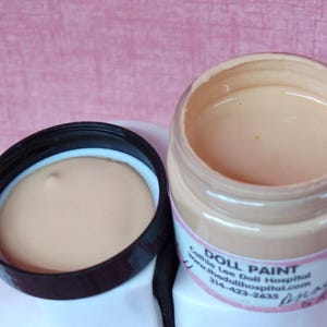 May include: An open jar of doll paint with a light peach color, next to its black lid. The jar has a white label with the text "DOLL PAINT" and contact information. The background is a pink surface.