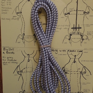 May include: A close-up of a white elastic cord with black stripes. The cord is coiled and ready to be used for stringing dolls. The image also includes a diagram of a doll with instructions for stringing it.