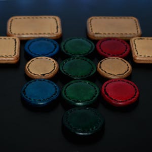 Op de afbeelding: Een assortiment handgemaakte lederen items in verschillende kleuren. De afbeelding toont twee rechthoekige en twee vierkante leren dozen met gestikte randen, samen met verschillende ronde leren stukken in blauw, groen en rood.