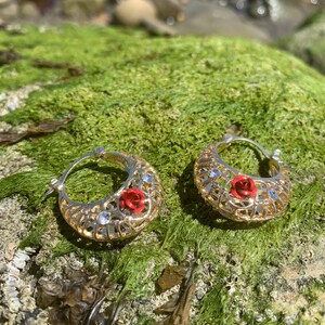 May include: A pair of gold hoop earrings with a delicate filigree design and red rose accents. The earrings are set against a backdrop of green moss.