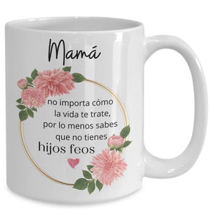 May include: White ceramic coffee mug with a gold rim and pink floral design. The mug has the text "Mam&aacute;" and the Spanish phrase "no importa c&oacute;mo la vida te trate, por lo menos sabes que no tienes hijos feos" in black text.