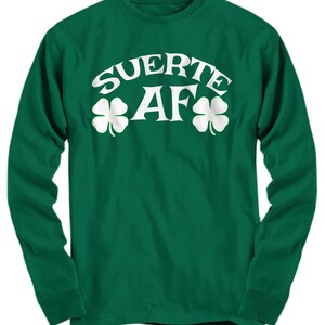 Könnte beinhalten: Grünes Langarmshirt mit weißem Text "SUERTE AF" und zwei Kleeblättern auf beiden Seiten des Textes.