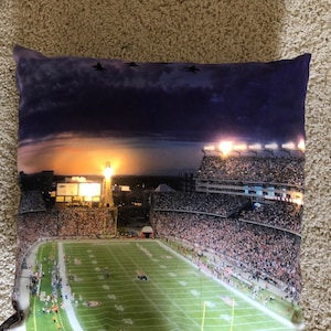 May include: A decorative pillow featuring a nighttime aerial view of a packed football stadium with a lit up scoreboard and a green field. The stadium is illuminated by bright lights against a purple and blue sky.