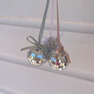 May include: Three silver disco ball ornaments with white and pink ribbon accents hanging from a white background.