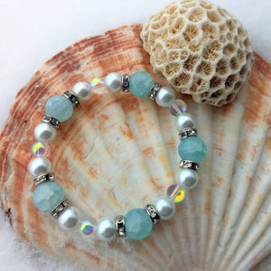 May include: A light blue and white beaded bracelet with clear iridescent beads and silver accents. The bracelet is displayed on a seashell.