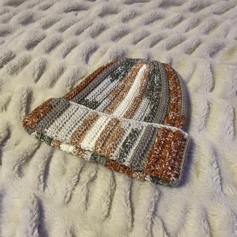 May include: A striped crocheted beanie hat in shades of brown, gray, and white. The hat is made with a soft, textured yarn and has a slouchy fit.