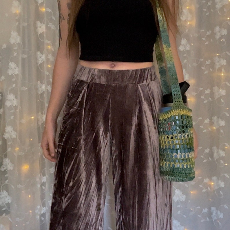 May include: A person wearing a black crop top and brown velvet pants with a crocheted green and blue bag slung over their shoulder.