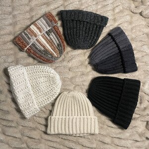May include: Six knitted hats in various colors and textures. The hats are arranged in a circle on a fuzzy white surface. The hats are all different styles, including a striped hat, a black hat, a white hat, and a gray hat.