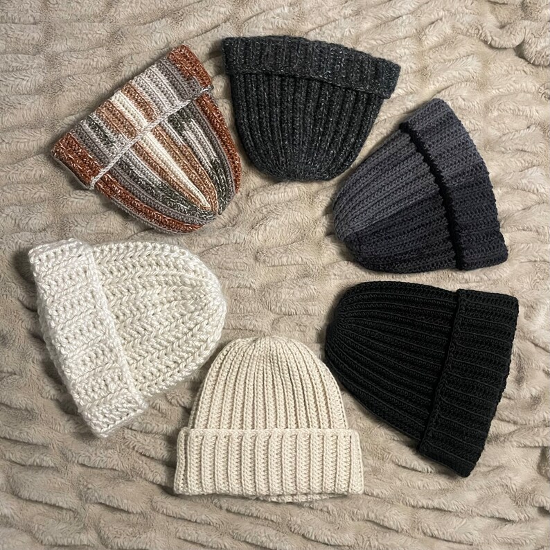 May include: Six knitted hats in various colors and textures. The hats are arranged in a circle on a fuzzy white surface. The hats are all different styles, including a striped hat, a black hat, a white hat, and a gray hat.
