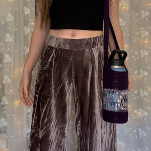 May include: A purple crocheted water bottle holder with a silver and blue design. The holder is worn as a crossbody bag and is being held by a person wearing brown velvet pants.