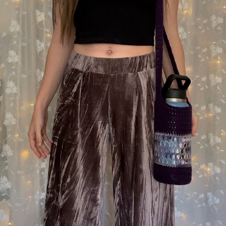 May include: A purple crocheted water bottle holder with a silver and blue design. The holder is worn as a crossbody bag and is being held by a person wearing brown velvet pants.