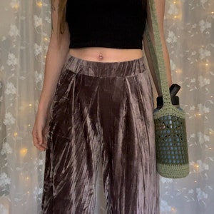 May include: A person wearing a black crop top and brown velvet pants with a crocheted green bag hanging from their shoulder. The bag is holding a blue water bottle.