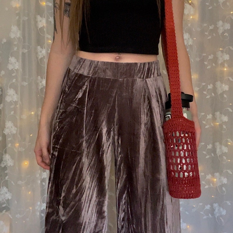 May include: A woman wearing a black top and brown velvet pants with a red crocheted bag hanging from her shoulder.