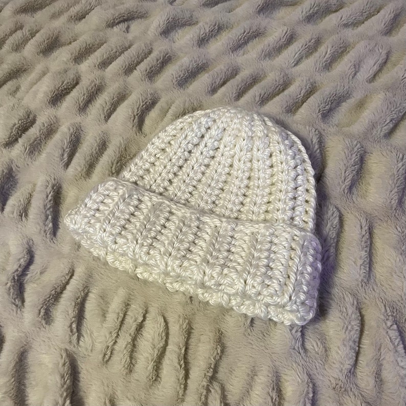 May include: A white crocheted beanie hat with a textured stitch pattern. The hat is folded down at the brim.