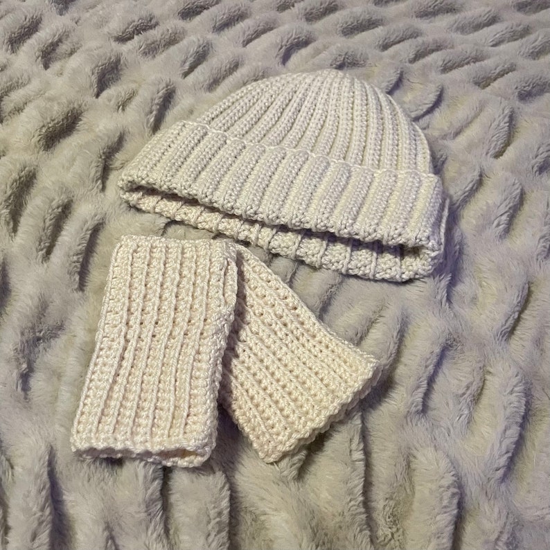 May include: A white crocheted beanie and matching wrist warmers. The beanie has a ribbed texture and the wrist warmers have a simple stitch pattern.