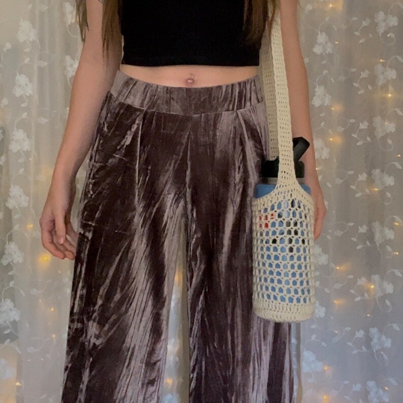 May include: A person wearing brown velvet pants and a black crop top is carrying a white crocheted bag with a blue water bottle inside.