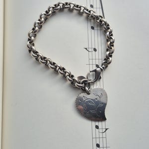 May include: A silver-toned bracelet with a heart-shaped charm. The charm features an engraved floral design and is attached to a chain-link bracelet with a lobster clasp. The bracelet is resting on a sheet of music.