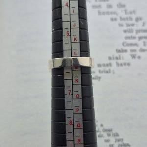 May include: A silver ring is being measured on a black ring sizer. The ring is placed on the size 6.5 mark. The background is a page of printed text.