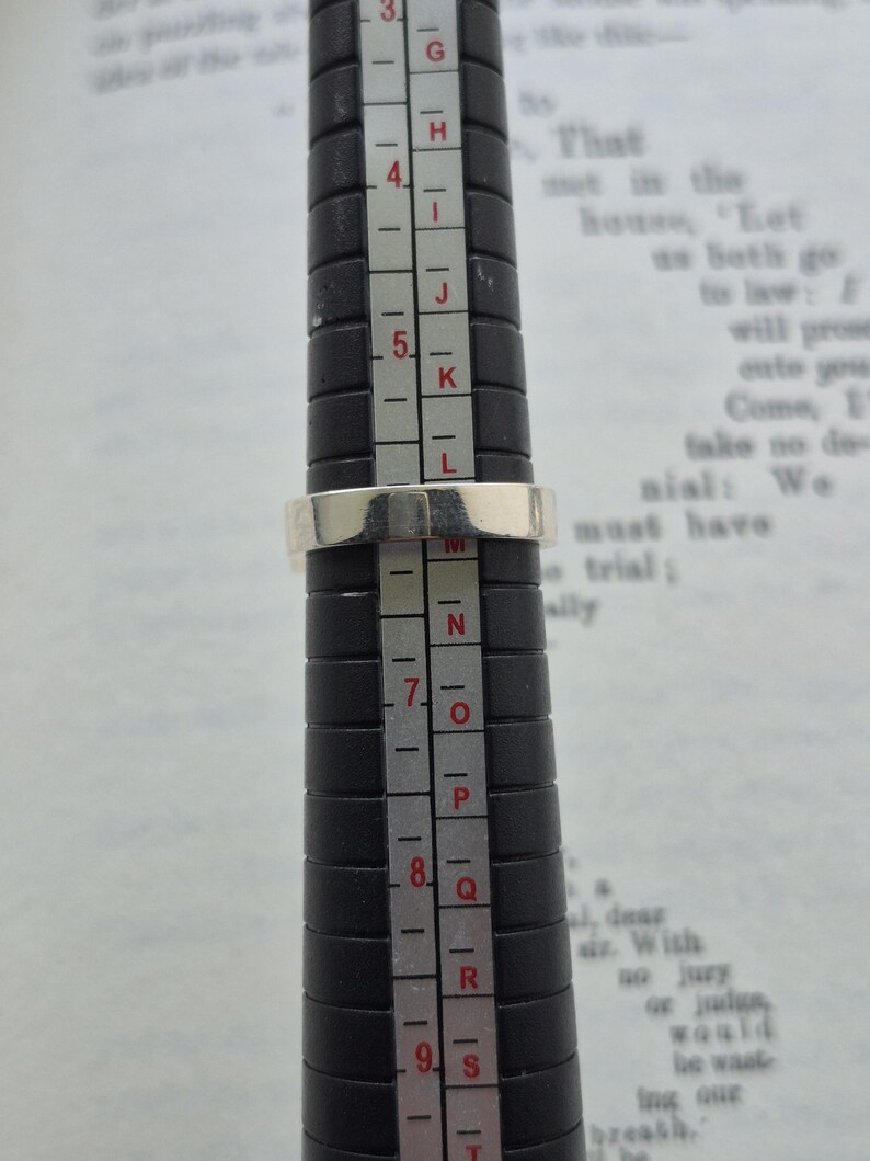 May include: A silver ring is being measured on a black ring sizer. The ring is placed on the size 6.5 mark. The background is a page of printed text.