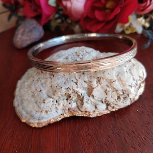 May include: A gold and silver bangle bracelet with a textured design. The bracelet is resting on a white, textured surface.