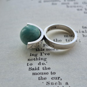 May include: A sterling silver ring featuring a teal-colored globe design. The ring has a split shank and a spherical teal stone. The ring is resting on a book page with printed text.