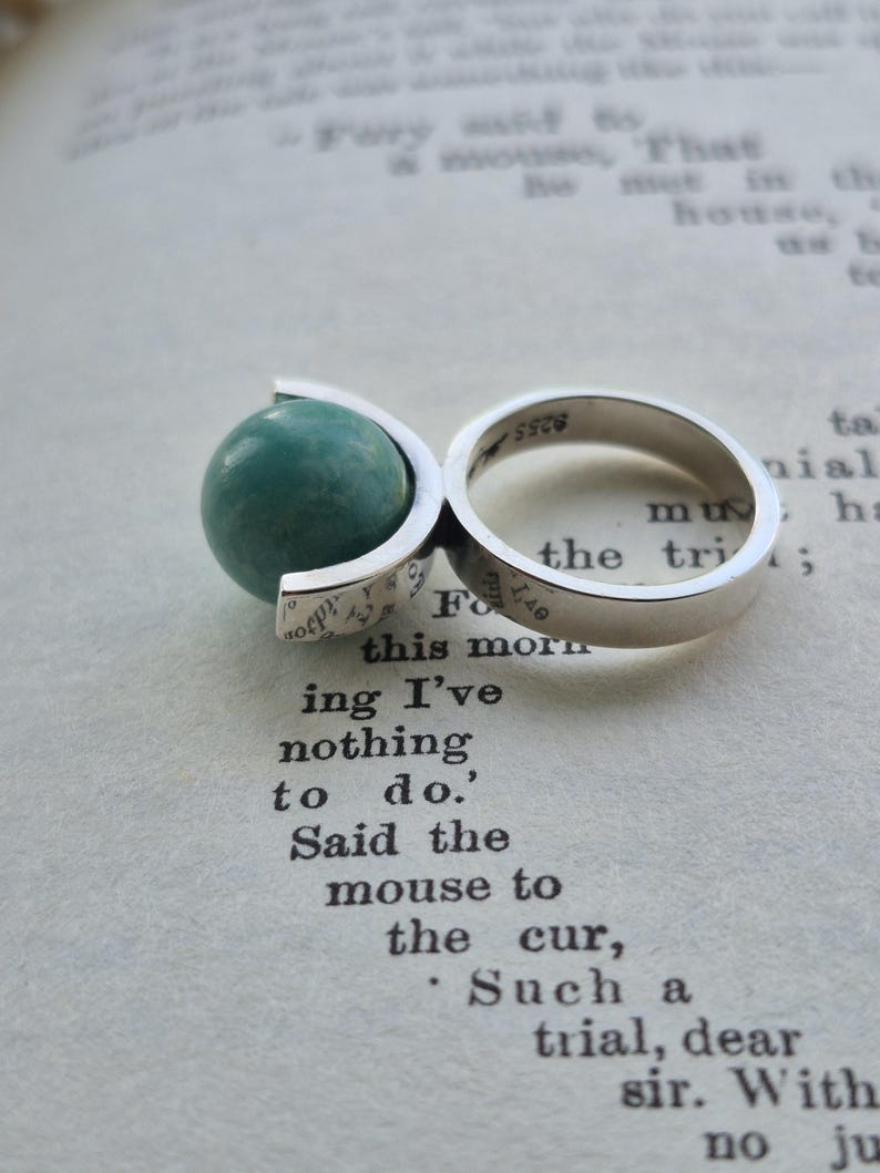 May include: A sterling silver ring featuring a teal-colored globe design. The ring has a split shank and a spherical teal stone. The ring is resting on a book page with printed text.