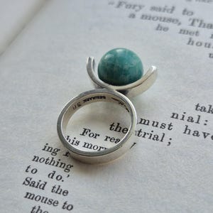 May include: A silver ring with a teal-colored, spherical stone. The ring's band is smooth and simple, with the word "DENMARK" stamped on the inside. The ring is set against the backdrop of an open book.