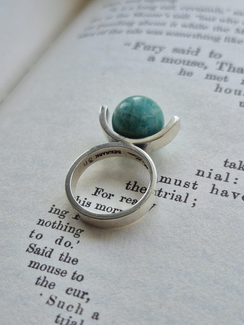 May include: A silver ring with a teal-colored, spherical stone. The ring's band is smooth and simple, with the word "DENMARK" stamped on the inside. The ring is set against the backdrop of an open book.