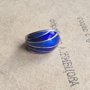 May include: A silver ring featuring a vibrant blue enamel inlay with a curved, segmented design. The surface of the blue enamel displays a fine, linear texture, while the silver bands create distinct, smooth, and polished metallic dividers.