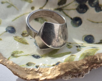 May include: A silver ring with a geometric design sits on a white ceramic dish with a gold rim and a floral pattern.