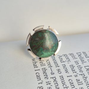 Op de afbeelding: Een zilveren ring met een grote, ronde, groen en bruine steen. De ring heeft een uniek ontwerp met zilveren accenten. De ring ligt op een open boek.