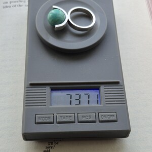 May include: A silver ring with a green stone is displayed on a digital scale. The scale reads 7.371. The ring has a unique design, with the stone held in place by a curved silver band. The scale is gray, and the background includes a book.