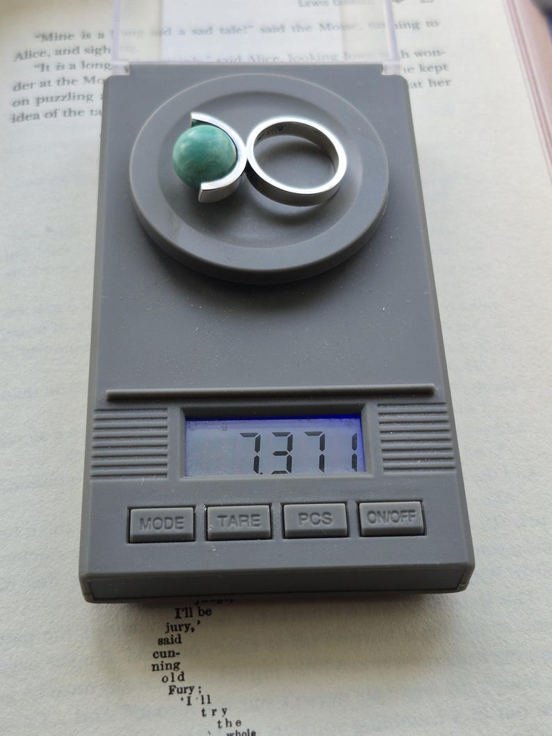 May include: A silver ring with a green stone is displayed on a digital scale. The scale reads 7.371. The ring has a unique design, with the stone held in place by a curved silver band. The scale is gray, and the background includes a book.