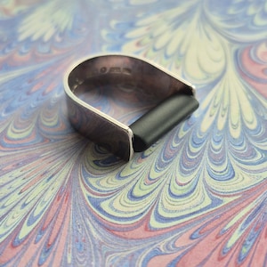 May include: A silver-toned ring with a black insert, resting on a colorful, patterned surface. The ring has a curved design with a rectangular black piece in the center. The background features a swirling pattern of pink, blue, and yellow.