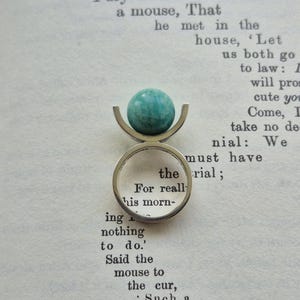 May include: A silver ring with a teal-colored, spherical stone. The ring's design features a curved, open setting that cradles the stone. The ring is set against a backdrop of open book pages.