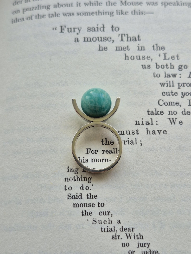 May include: A silver ring with a teal-colored, spherical stone. The ring's design features a curved, open setting that cradles the stone. The ring is set against a backdrop of open book pages.