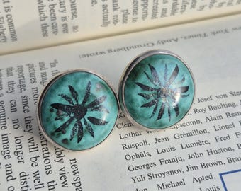 Könnte beinhalten: Paar runde, türkisfarbene Ohrringe mit silbernem Rand. Jeder Ohrring hat ein schwarzes, stilisiertes Blumendesign. Die Ohrringe liegen auf einem offenen Buch.