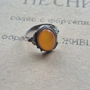 May include: A silver ring featuring an oval-shaped, smooth, translucent orange stone set in a decorative metal frame with small bead-like accents along the sides of the setting, resting on a textured, light-colored surface with printed text.