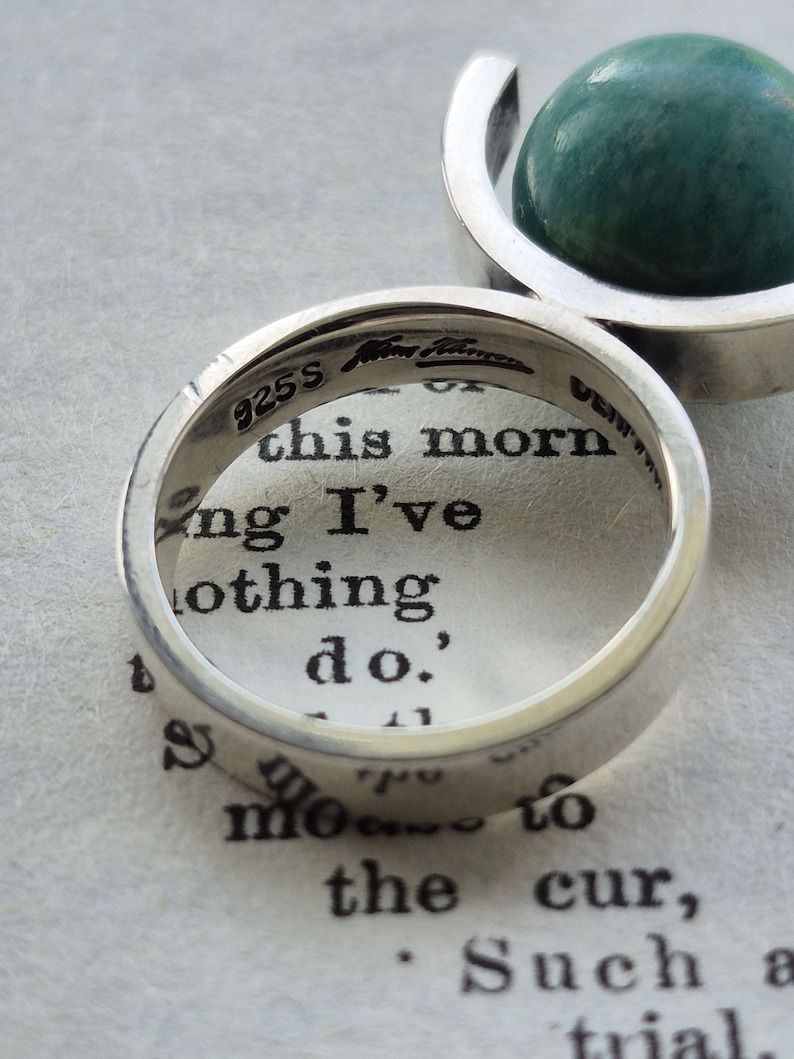 May include: Close-up of two silver rings. One ring is in focus, displaying the text "925S" and other markings. The second ring holds a green, spherical stone. The rings are on a page with printed text.