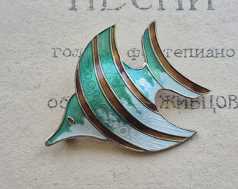 May include: A vintage fish-shaped brooch with a detailed design. The brooch features a combination of teal, brown, and white enamel, with a silver-tone metal outline. The design includes circular and linear patterns, creating a textured appearance. The brooch is set against a backdrop of aged paper.