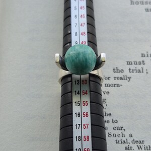 May include: A silver ring with a large, round, green gemstone. The ring is on a black ring sizer, with measurements in inches and millimeters. The ring's design features a modern, minimalist style, with the gemstone as the focal point.