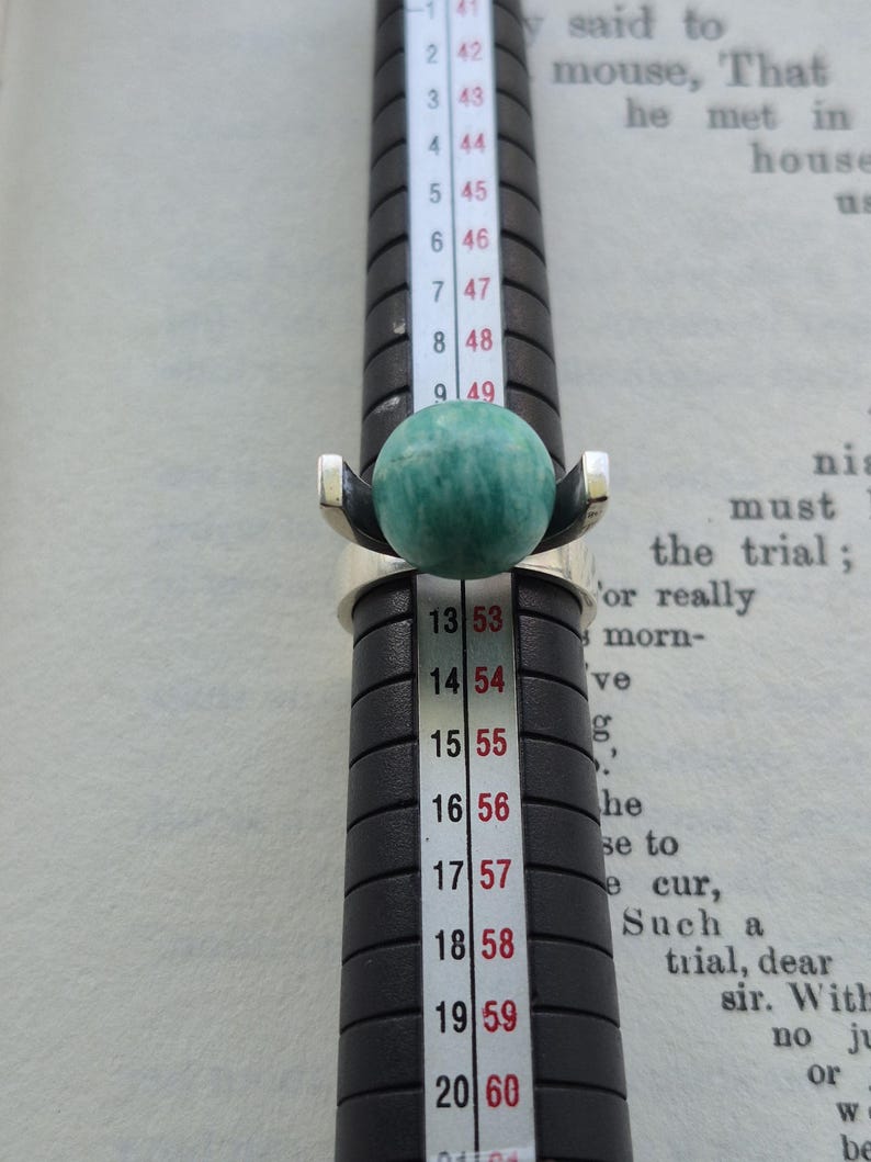 May include: A silver ring with a large, round, green gemstone. The ring is on a black ring sizer, with measurements in inches and millimeters. The ring's design features a modern, minimalist style, with the gemstone as the focal point.