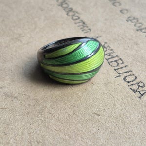 May include: A dark metal ring featuring a series of curved, raised segments filled with vibrant lime and emerald green enamel. The surface of the enamel sections displays fine, linear textures, while the metal frame remains dark and smooth.