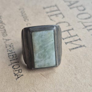 May include: A rectangular silver ring with a light green stone inlay. The ring has a thick band and a rectangular face with a pale green stone. The ring appears to be vintage or antique.