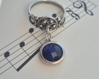May include: A silver ring featuring an intricate band design and a hanging, circular, deep blue gemstone. The ring is displayed on a background of musical notation, possibly indicating a musical or artistic theme. The gemstone is about 1.3 cm in diameter.