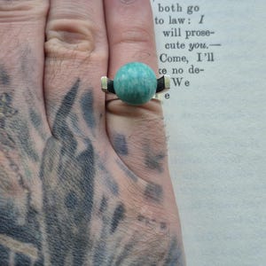 May include: A silver ring with a large, round, turquoise-colored stone. The ring has a modern design with black and gold accents on the band. The ring is shown on a hand.