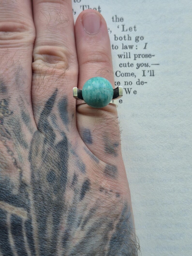May include: A silver ring with a large, round, turquoise-colored stone. The ring has a modern design with black and gold accents on the band. The ring is shown on a hand.