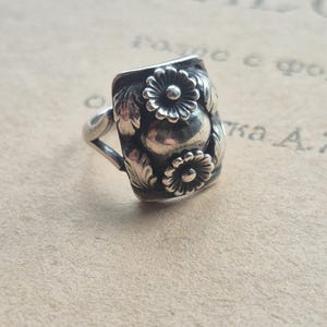 Puede incluir: Anillo de plata con una cara rectangular con un diseño floral. El anillo tiene dos flores y hojas, con una apariencia antigua detallada. La banda es simple y delgada, sobre un fondo neutro.